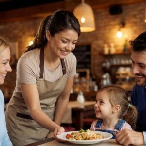 A smiling restaurant server serving food to a happy child and her parents.